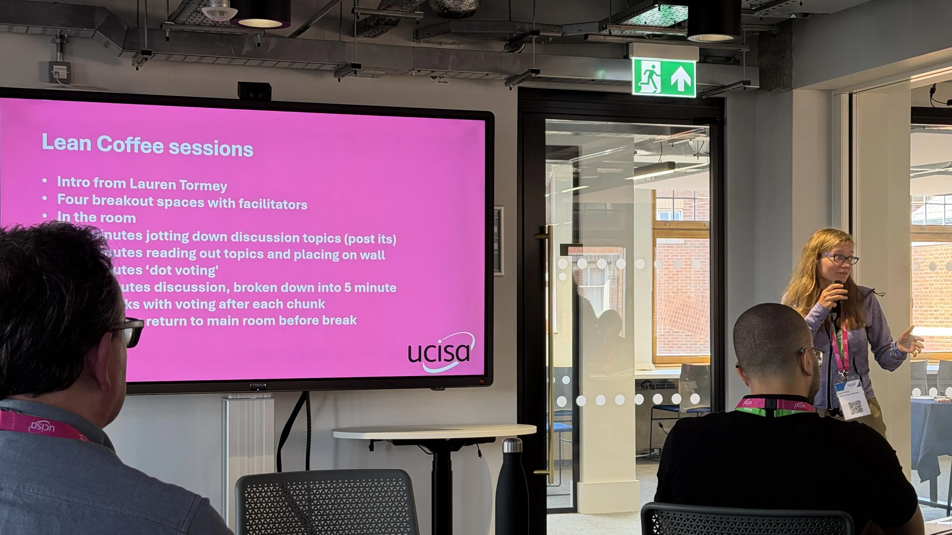A Lauren Tormey speaks to an audience in a modern room with a large pink slide titled Lean Coffee Sessions.