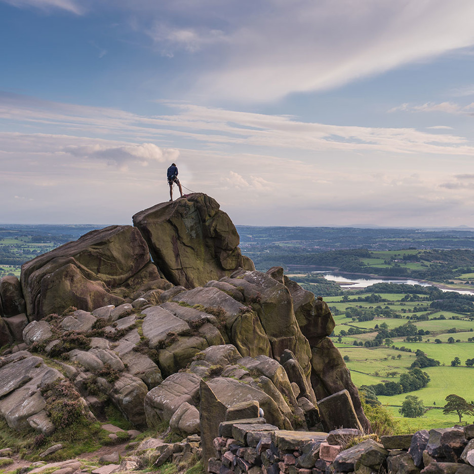 A rock climber standing at the summit overlooking a green valley.