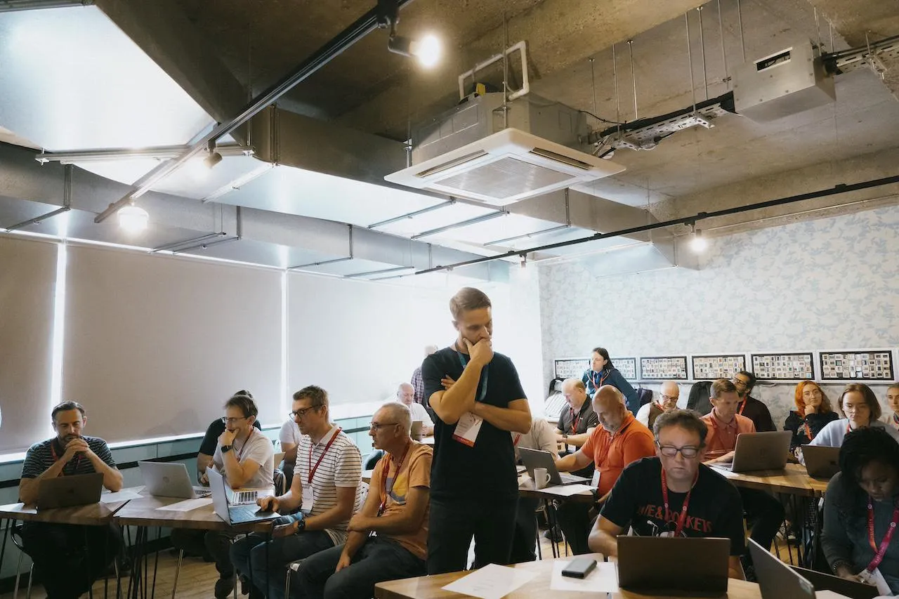 A man standing looking thoughtfully at a computer screen, with people sitting at laptops in the background.